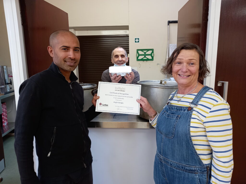 Sue, woman with dark hair, stripy t-shirt and dungarees, is giving a certificate to two owners of a local Bangladeshi restaurant, both men dressed in dark colours in a kitchen