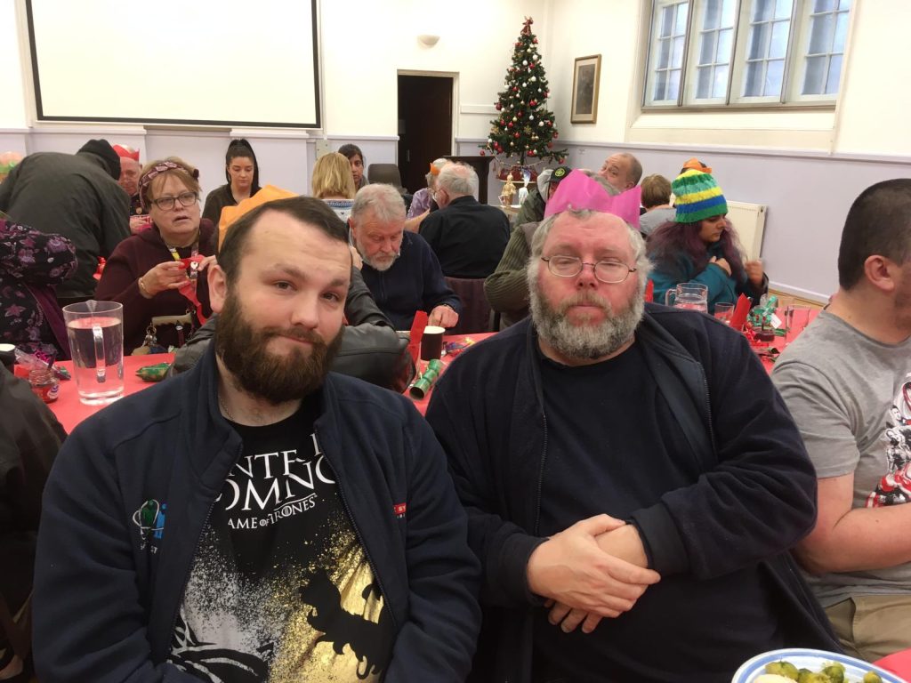 people sitting around tables with a christmas tree in the background. Two men with beards sitting in the foreground facing the camera