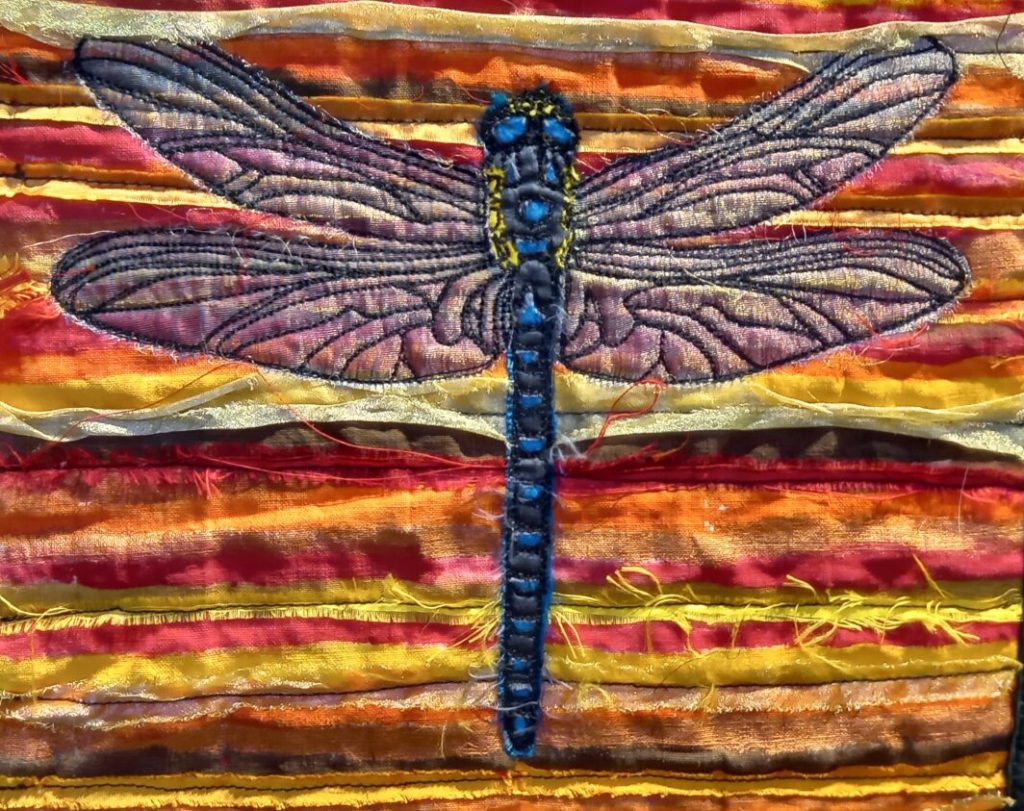 A picture of part of a textile art piece showing a stitched dragonfly on a multicoloured striped background